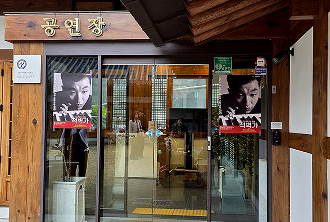 서울돈화문국악당 ‘공연장’ 입구. 유리문 양쪽에 흑백 인물 사진이 담긴 공연 포스터 ‘적벽가’가 붙어 있으며, 안쪽에는 안내 데스크와 관람객 몇 명이 보인다. 나무 기둥과 전통창호가 어우러진 한옥 같은 건물 외관이 특징적이다.