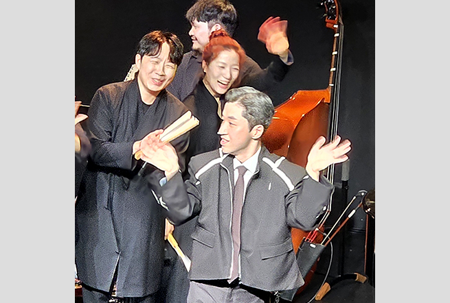 커튼콜 중 네 명의 연주자가 손을 흔들며 인사하고 있다. 앞에는 은색으로 염색한 머리와 정장을 입은 최준 음악가가, 뒤편에는 검은 의상을 입은 다른 연주자들이 미소 지으며 서 있다. 오른쪽에는 콘트라베이스가 세워져 있다.