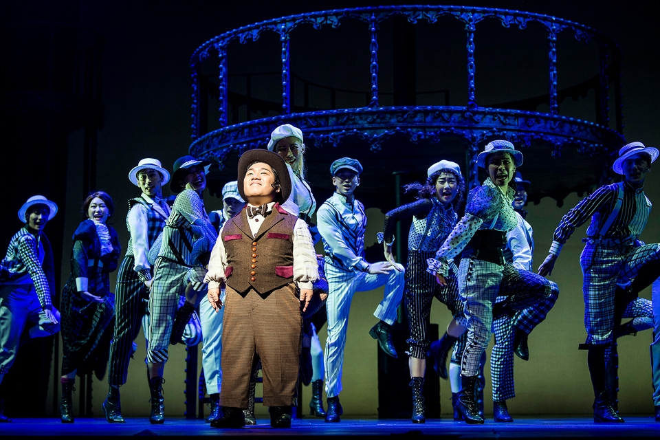 무대 중앙에 김유남 배우가 정면을 바라보고 서 있다. 조끼와 와이셔츠, 보타이를 갖춘 클래식한 의상에 모자를 썼다. 뒤에는 앙상블 배우들이 나란히 서서 군무 동작을 취하고 있다.