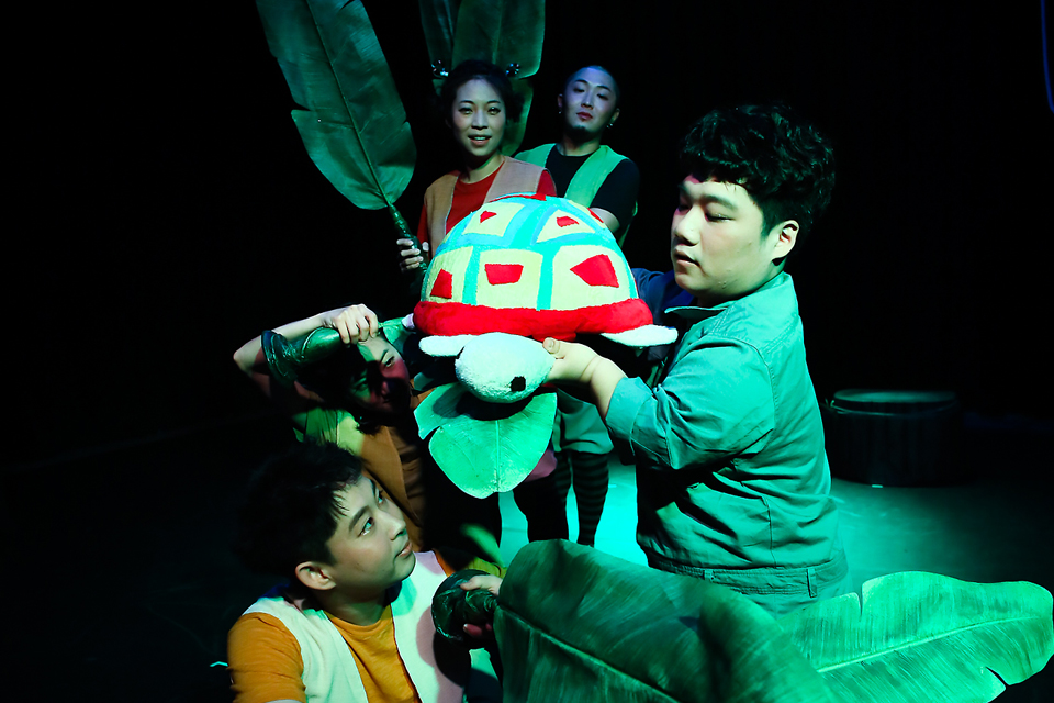 김유남 배우가 알록달록한 등딱지를 가진 거북이 인형을 두 손으로 들어 올리고 있고, 주변의 배우들은 큰 잎사귀 모양 소품을 들고 거북이를 둘러싸듯 서 있다. 초록빛 조명이 자연 속 분위기를 자아낸다.