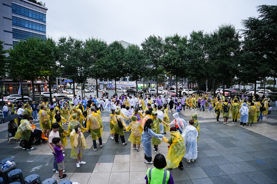 도심 광장에서 노란색과 하늘색 우비를 입은 참가자 수백 명이 원을 이루며 서로 손을 맞잡고 대동놀이를 즐기고 있다.