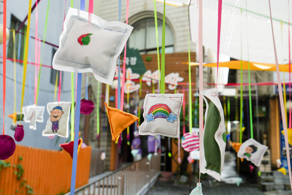 소극장 함세상 앞마당에 알록달록한 리본과 천이 길게 드리워져 있고, 사람 얼굴, 무지개, 하트 등을 수놓은 작은 천 쿠션 장식들이 공중에 매달려 있다.