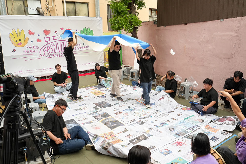 마당에 신문지가 넓게 깔려 있고, 검은 티셔츠를 맞춰 입은 배우들이 신문지 경계선을 따라 앉아 있다. 세 사람이 긴 천을 맞잡아 머리 위로 높이 들고 신문지 위를 걷고 있다.