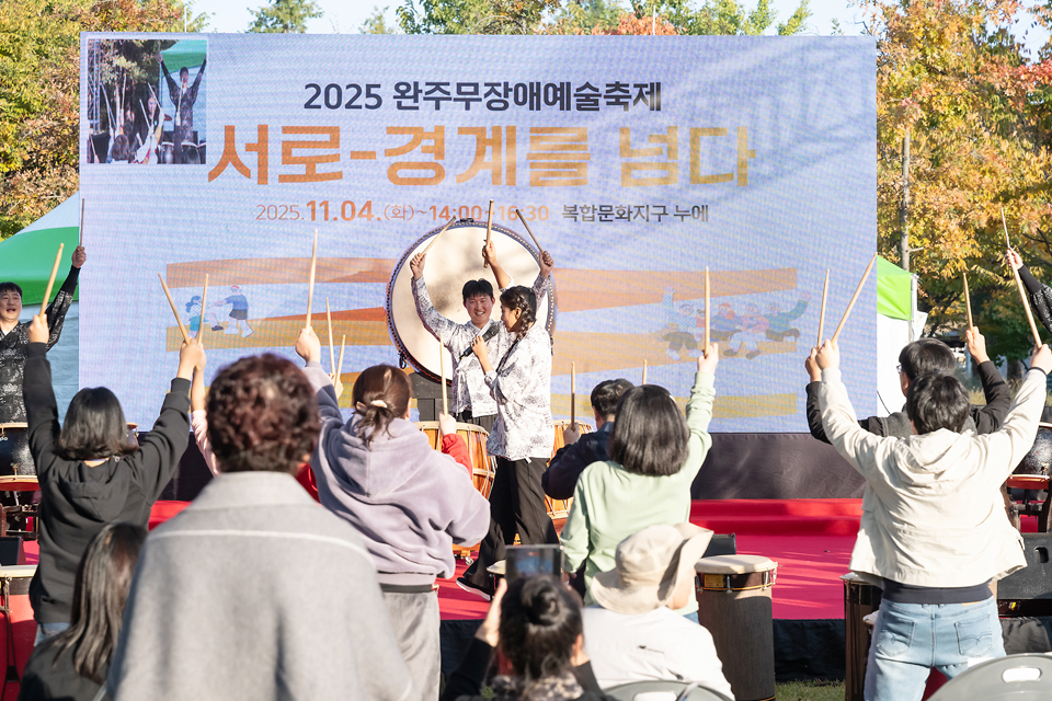 야외무대 중앙에 놓인 큰북 앞에서 연주자가 양손에 북채를 들고 관객을 향해 서 있고, 관객들이 무대의 연주자를 바라보며 양손에 북채를 들고 신나 한다. 무대 백드롭에는 ‘2025 완주무장애예술축제 서로-경계를 넘다’라고 쓰여 있다.