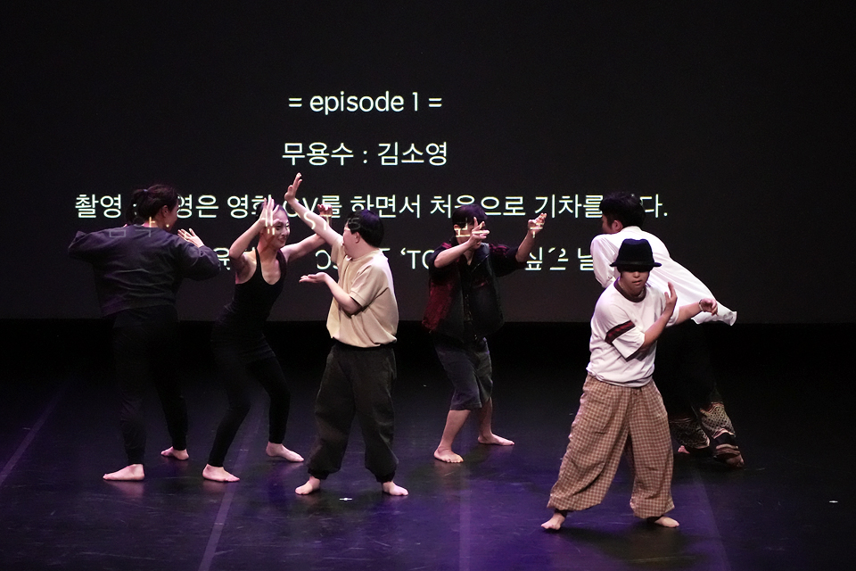 여섯 명의 무용수가 어두운 무대 위에서 서로 손을 맞잡거나 교차시키며 역동적인 동작을 펼치고 있다. 뒤편 스크린에는 ‘episode 1’, ‘무용수: 김소영’ 등의 글자가 자막으로 비춘다.