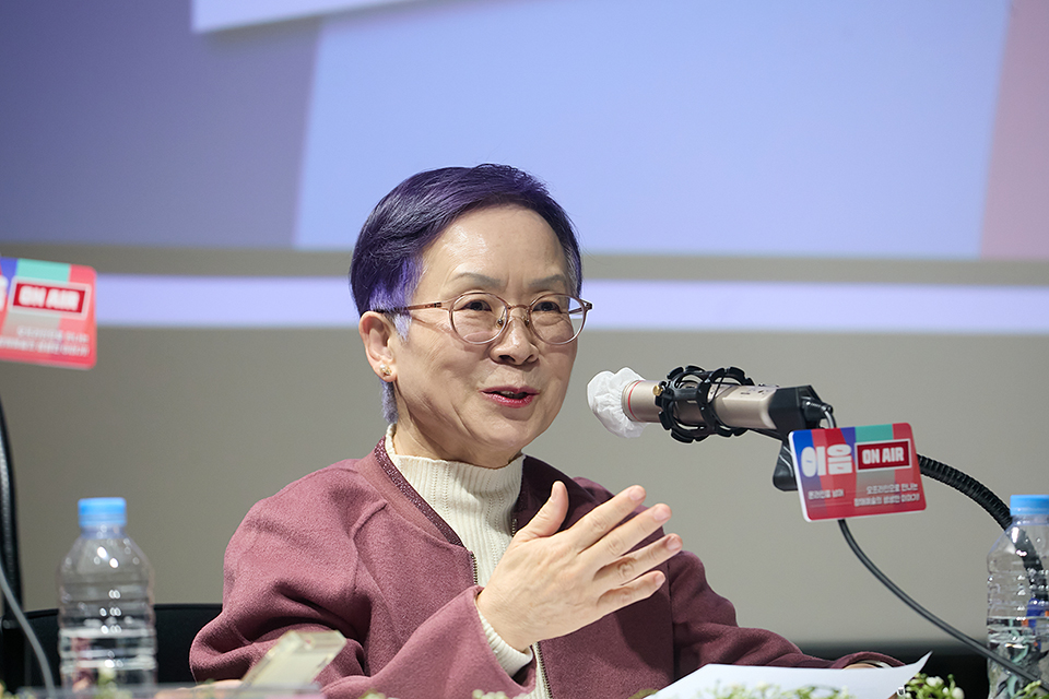 보라색으로 염색한 짧은 머리에 짙은 분홍색 재킷을 입은 작가가 마이크 앞에 앉아 말하고 있다.