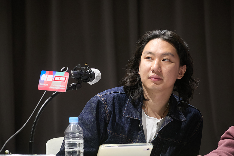 긴 머리에 청재킷을 입은 채 마이크 앞에 앉아 말하고 있다.