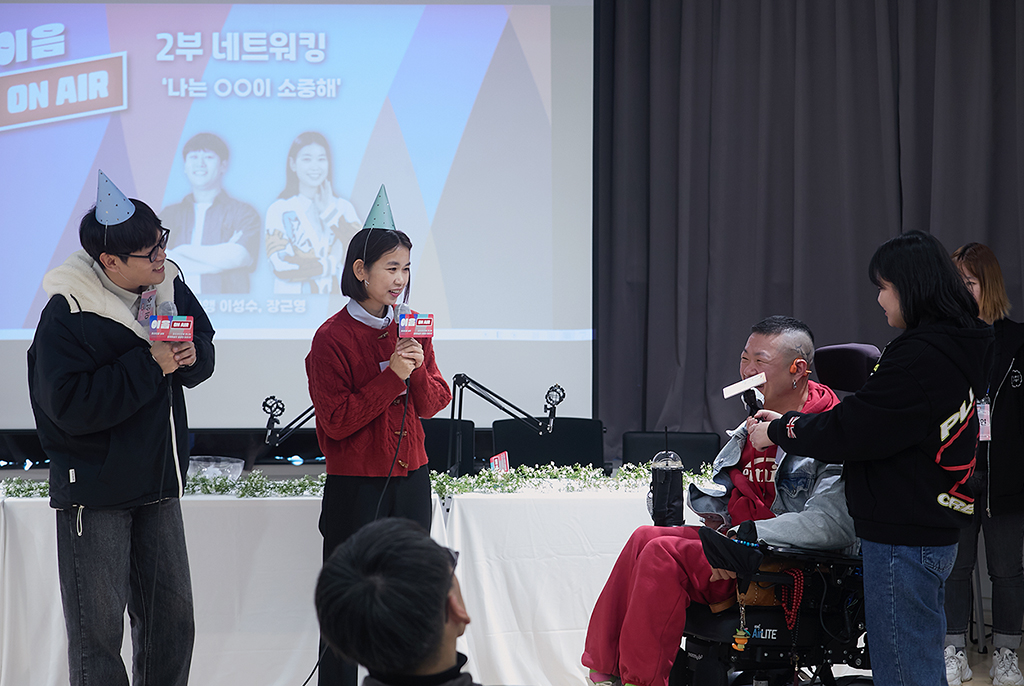 무대 앞에서 고깔모자를 쓴 진행자 이성수 대표와 장근영 작가가 마이크를 들고서 전동휠체어를 이용하는 참가자와 이야기를 주고받고 있다. 뒤쪽으로 영상 스크린에는 ‘이음온에어 2부 네트워킹. 나는 ○○이 소중해’라는 글과 함께 두 진행자 사진이 있다.