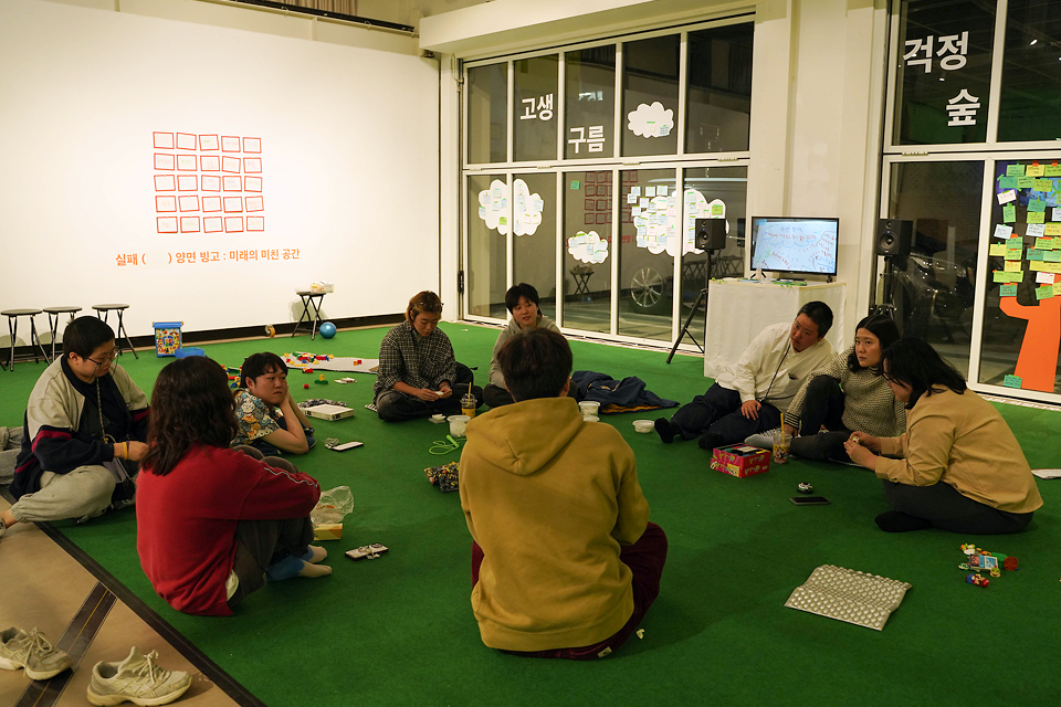 초록색 바닥 매트 위에 둥글게 앉아 있다. 따뜻한 조명이 비추고, 주변에는 메모지와 필기도구, 소품들이 놓여 있다. 커다란 유리벽에는 걱정, 숲, 고생, 구름 등의 글자와 여러 색의 포스트잇이 붙어 있다.