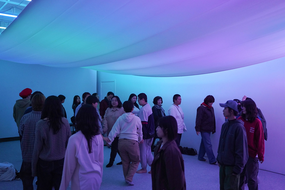 푸른빛과 보랏빛이 섞인 은은한 조명의 체험 공간에서 여러 참가자들이 자유롭게 거닐고 있다. 천장은 부드러운 천으로 덮여 파도처럼 굽이치며 빛을 반사한다.