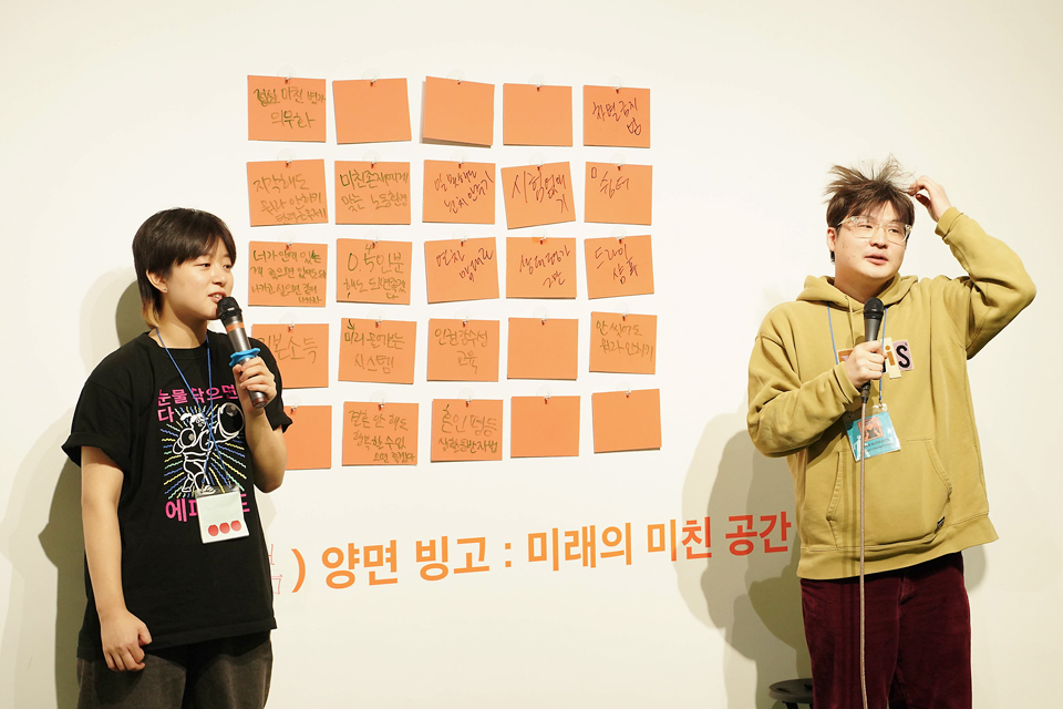 흰 벽에 주황색 메모지가 격자 형태로 붙어 있고, 하단에 ‘양면 빙고: 미래의 미친 공간’이라고 쓰여 있다. 좌우에서 두 진행자가 마이크를 들고 관객과 소통하고 있다. 