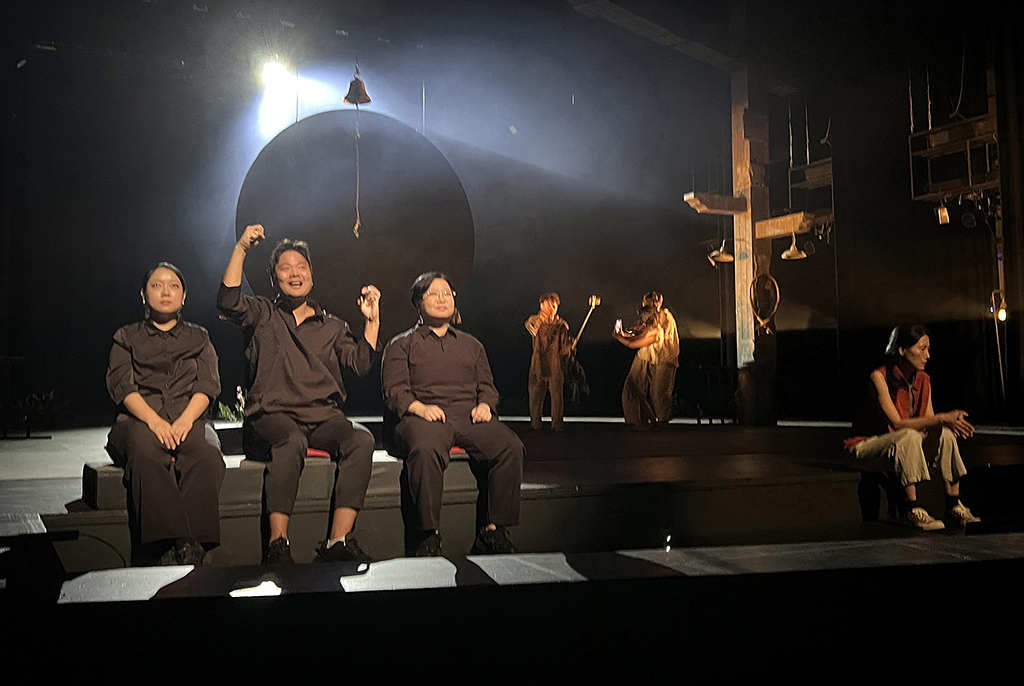 어두운 무대 한가운데 둥근 조명이 비추는 가운데, 세 명의 수어통역사가 검은 옷을 입고 왼쪽 앞에 나란히 앉아 있다. 가운데 통역사는 두 손을 들고 표정을 살리며 수어를 전달하고 있다. 무대 오른쪽 뒤에는 배우들이 서로 마주 보고 연기 중이며, 무대 앞 오른쪽 가장자리에는 한 배우가 앉아 있다.