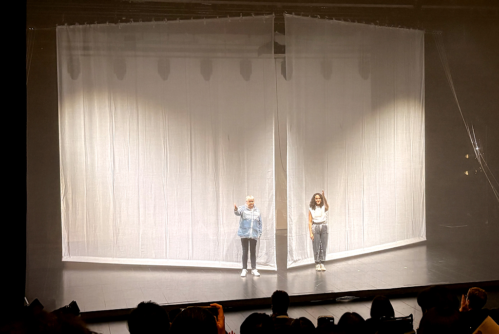 무대 위에 반투명한 흰 천으로 둘러싸인 사각 구조물이 설치되어 있고, 그 앞에 두 명의 출연자가 나란히 서서 관객을 향해 인사하고 있다.