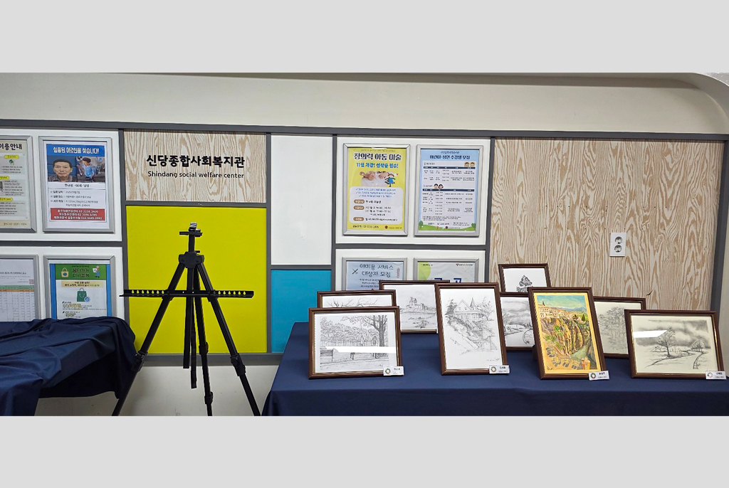 전시장 한쪽 벽면과 테이블 위에 자료와 작품이 함께 전시되어 있다. 벽에는 포스터와 안내문이 붙어 있고, 테이블 위에는 액자에 담긴 드로잉과 인쇄물이 펼쳐져 있으며, 중앙에는 이젤이 놓여 있다.