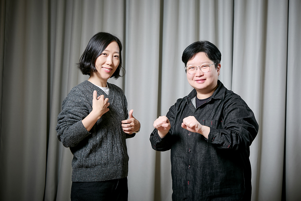 부드럽게 주름이 잡힌 회색 커튼을 배경으로 조희경, 지혜원 두 사람이 나란히 서서 카메라를 향해 미소 짓고 있다. 두 사람은 양손을 들어 수어의 [반갑다] [안녕] 의미를 연상시키는 동작을 하고 있다.