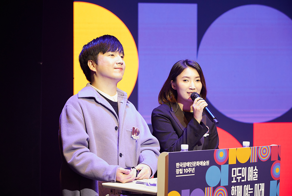 신홍윤 강사는 연보랏빛 재킷을 입고 앞을 바라보며 미소 짓고 있고, 허우령 아나운서는 검은 재킷을 입고 마이크를 들고 발언 중이다. 두 사람 앞에는 ‘모두의 예술, 함께 여는 미래’라는 문구가 적힌 단상이 놓여 있다.