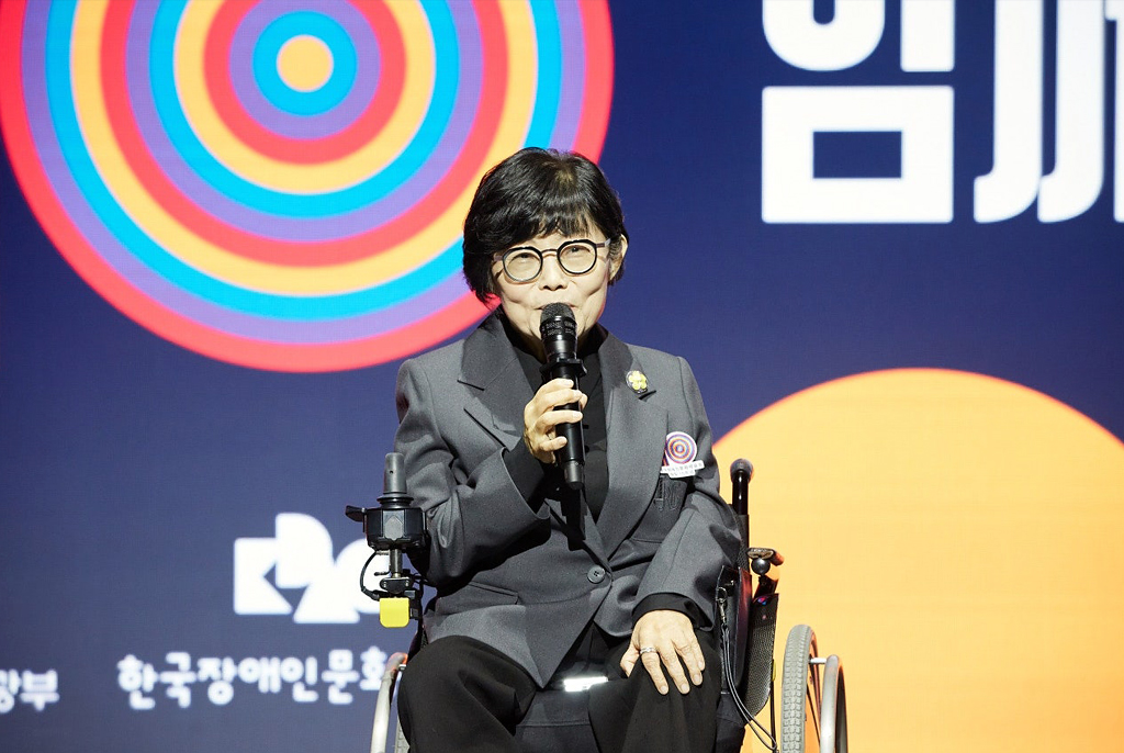 회색 정장을 입은 방귀희 이사장이 휠체어에 앉아 마이크를 들고 단정한 표정으로 관객을 향해 발언 중이다. 