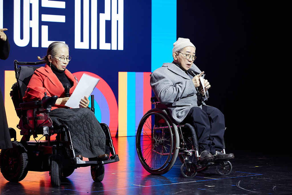 무대 중앙에 휠체어를 탄 두 사람이 있다. 이정희 자수장은 붉은색 상의에 검정 치마를 입고 손에 대본을 들고 있고, 고정욱 작가는 회색 코트와 하얀 비니를 착용한 채 마이크를 들고 발언 중이다. 