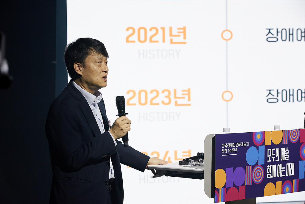 검정색 정장 차림의 조현성 위원이 무대 단상 앞에서 마이크를 들고 발표하고 있다. 뒤편 스크린에는 ‘2021년’, ‘2023년’ 등 연도가 표시된 장문원의 주요 연혁이 투사되고 있다.