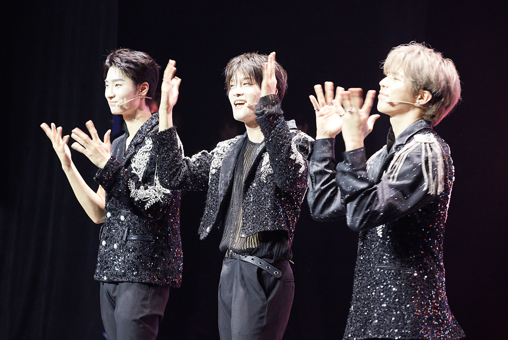 세 명의 멤버가 반짝이는 장식이 있는 검은색 의상을 입고 양 손을 얼굴 높이로 들어 수어로 노래하고 있다.