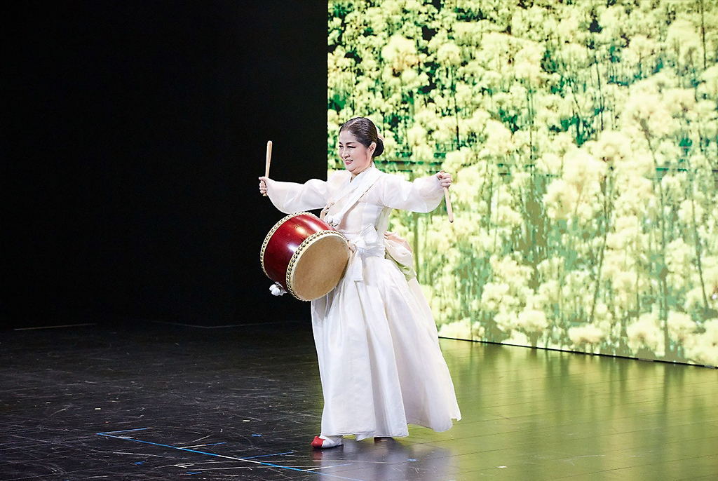 김영민 무용가가 쪽진 머리에 흰색 한복을 입고, 북을 어깨띠로 매고 춤을 추고 있다. 한 손에는 북채를 들고, 다른 손은 펼쳐 우아한 동작을 표현한다. 뒤편 스크린에는 하얀색 꽃밭 풍경이 크게 비추고 있다.