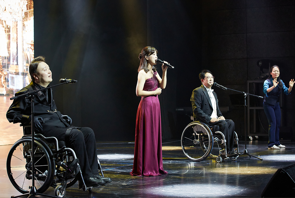 자주색 드레스를 입고 가운데에서 선 강유경 성악가의 왼편과 오른편에 검정색 정장을 입고 휠체어를 탄 이남현 성악가와 황영택 성악가가 노래하고 있다. 무대 오른편에는 수어통역사가 가사를 수어로 통역하고 있다.
