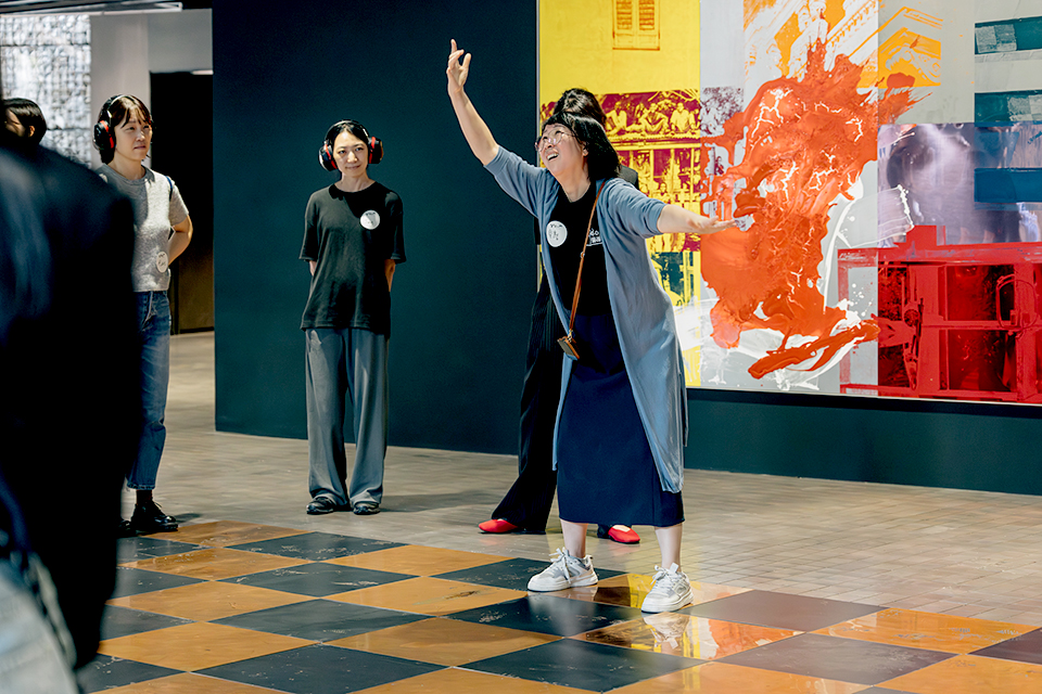 전시장 가운데에서 한 참여자가 한 손을 높이 들고 퍼포먼스를 펼치고 있고, 다른 참여자가 헤드셋을 착용한 채 이를 지켜보고 있다. 배경에는 강렬한 색감의 작품이 걸려 있어 생동감 있는 분위기다.