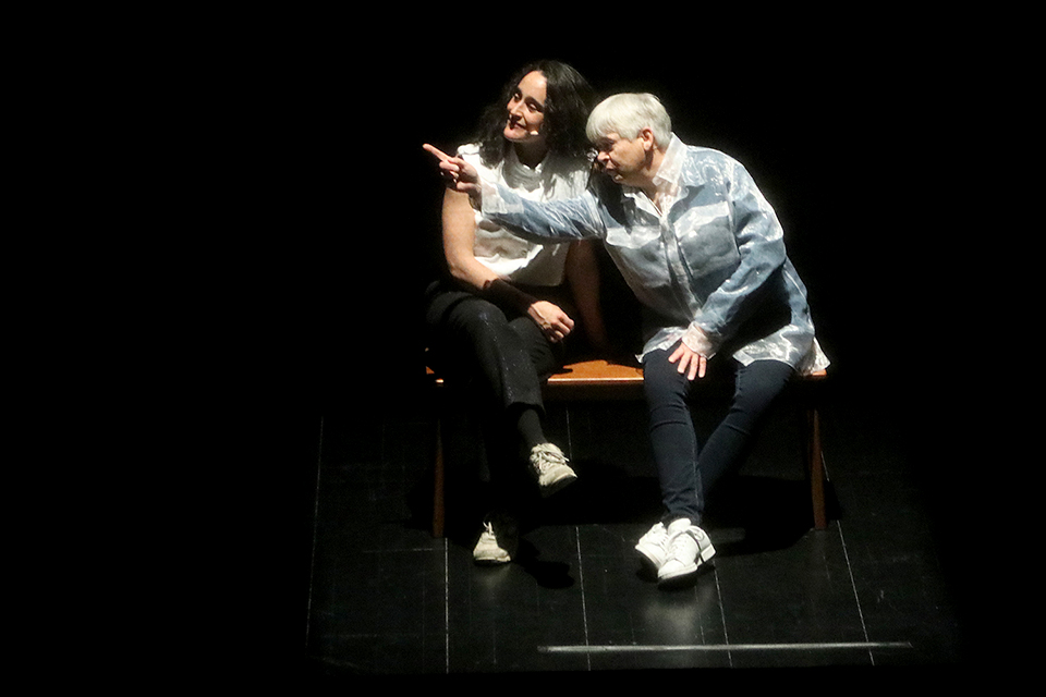 어두운 무대 위에 에바나와 힐다가 나란히 벤치에 앉아 있다. 힐다가 팔을 뻗어 무대 하수 어딘가를 가리키고 있고, 에바나는 힐다가 가리킨 쪽을 바라보면 미소 짓고 있다.