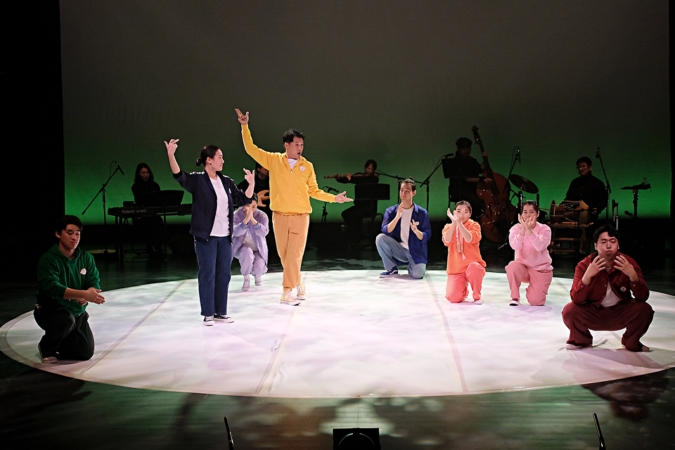 큰 원으로 비추는 조명 아래 빨주노초파남보 색깔의 트레이닝복을 입은 여덟 명의 출연자가 서거나 앉거나 몸을 낮춘 채 다른 동작으로 움직이고 있다. 뒤쪽에는 희미한 조명 아래 악사들의 실루엣이 보인다.