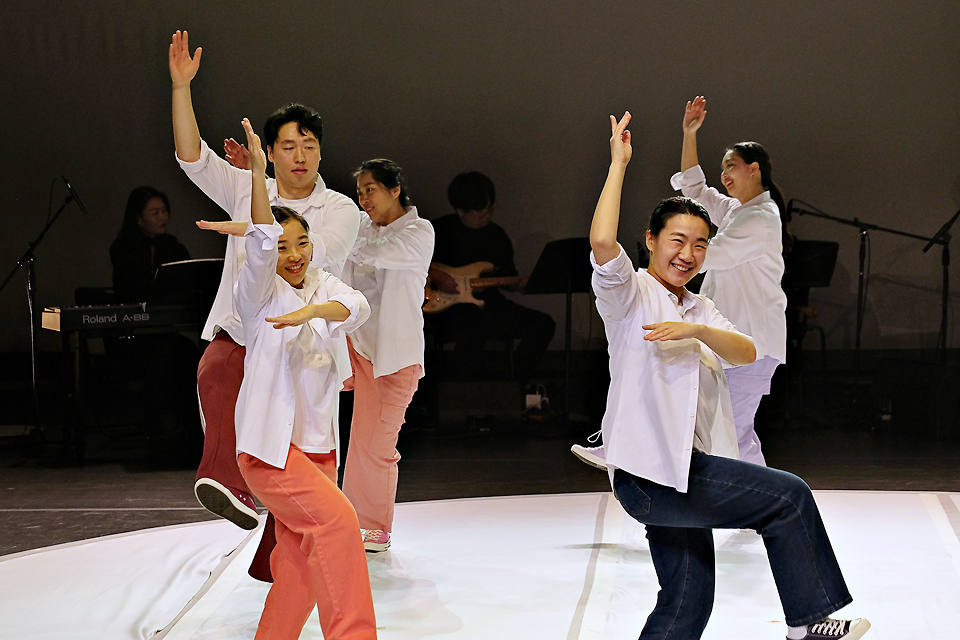 무대 위에서 하얀 셔츠로 갈아입은 여러 명의 출연자가 왼팔은 얼굴 아래에 가로로 놓고 오른팔은 머리 위로 높이 든 채, 한 발로 겅중 뛰며 탈춤을 추고 있다.