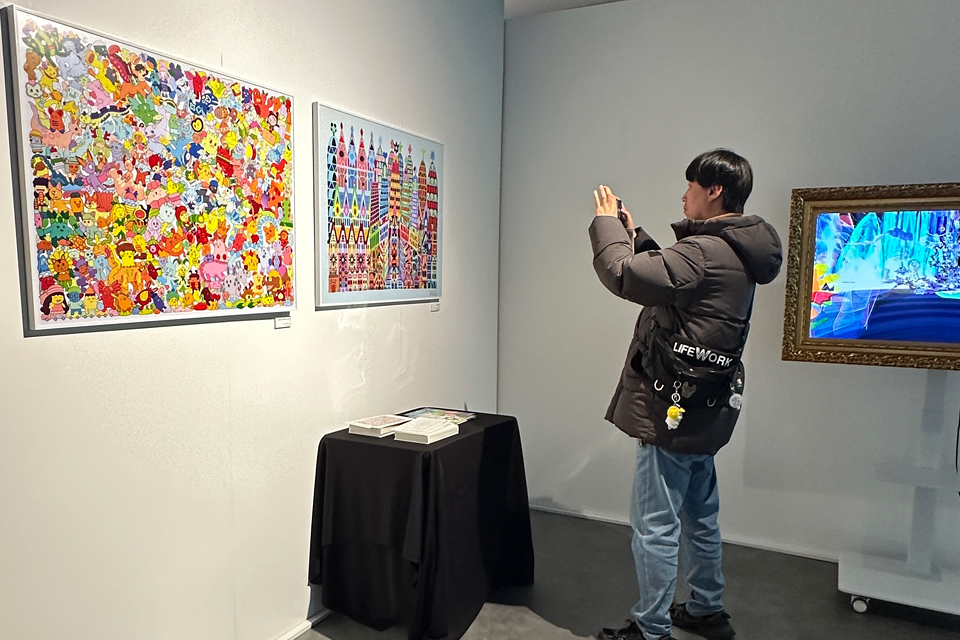 병철이 스마트폰으로 전시장 한쪽 벽에 걸린 신현채 작가의 회화 작품을 촬영하고 있다. 옆 벽면에는 소형 모니터가 디지털 액자처럼 걸려 있다.