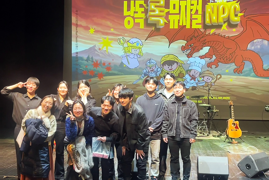 무대 앞에서 공연 참여자와 관객 10여 명이 모여 기념사진을 찍고 있다. 뒤쪽 대형 스크린에는 게임 일러스트 배경 위로 “낭독 록 뮤지컬 NPC”라고 공연 제목이 쓰여 있다. 스크린 앞으로 기타와 드럼 등 악기가 놓여 있다.