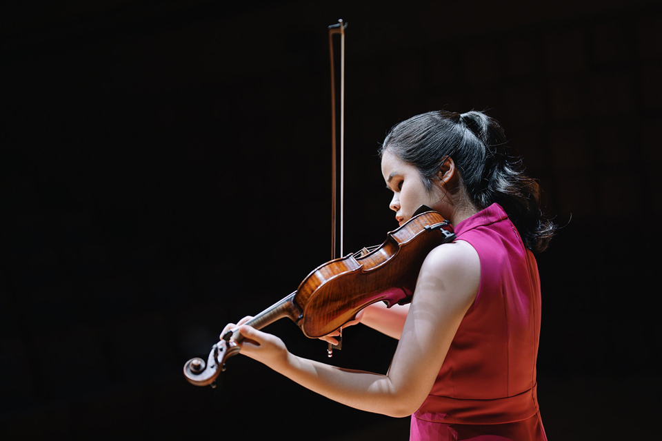 김지선 바이올리니스트가 검은 배경의 무대에서 붉은색 민소매 의상을 입고 바이올린을 연주하고 있다.
