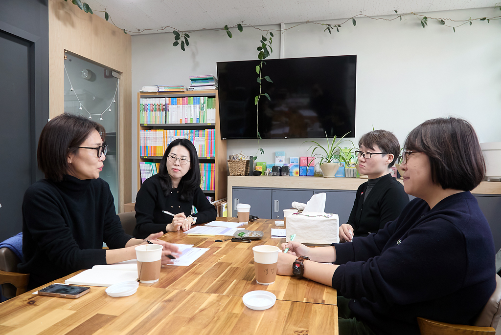 네 사람이 긴 테이블에 마주 앉아 서로를 바라보며 이야기 나누고 있다. 테이블 위에는 차와 자료가 놓여 있고, 정면으로 벽에는 모니터가 매달려 있고, 책이 가득 꽂힌 책장이 있고, 모니터 아래 선반에는 작은 교구와 화분들이 놓여 있다.