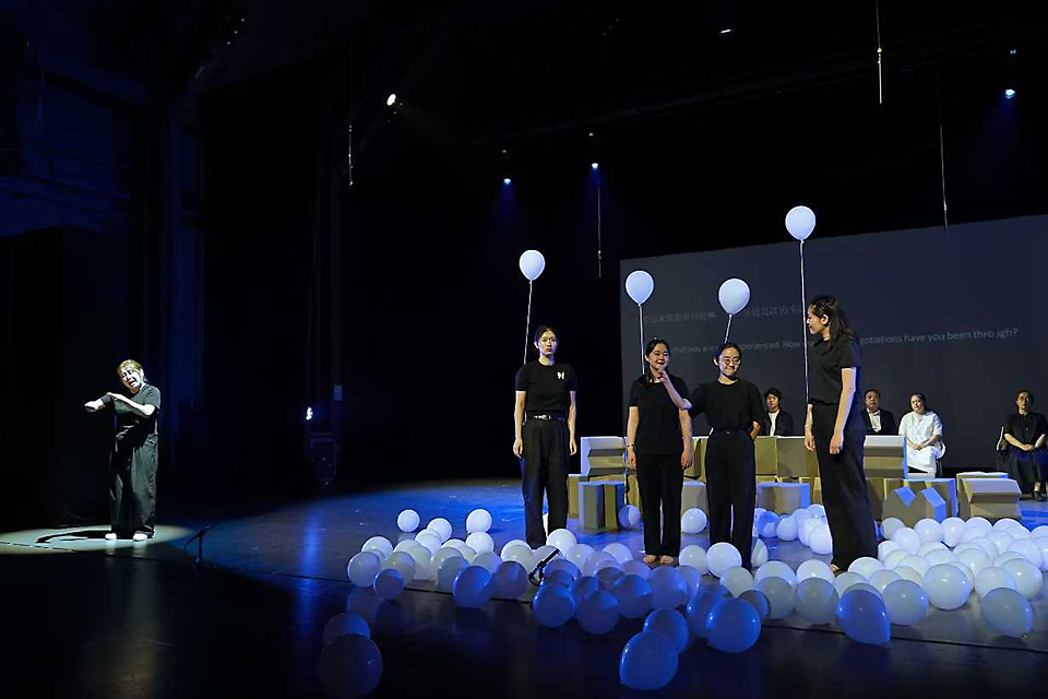 풍선을 머리 위로 달고 검은 옷을 입은 배우 네 명이 나란히 서있고, 바닥에는 풍선이 가득 놓여 있다. 뒤편으로 배우들이 나란히 책상을 두고 앉아 있다. 무대 왼편에서는 수어통역을 하고 있다.