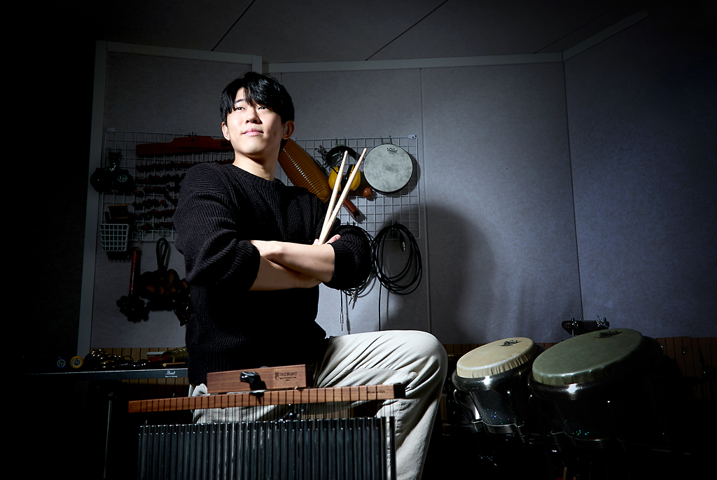 다양한 브라질 악기로 채워진 작업실에서, 이성재 퍼커셔니스트가 팔짱을 낀 채 한 손에 드럼 스틱을 들고 살짝 고개를 위로 향해 있다. 앞쪽으로 윈드차임과 봉고가 나란히 놓여 있고, 뒤편 벽에는 작은 손북 등 다양한 소형 타악기가 걸려 있다.