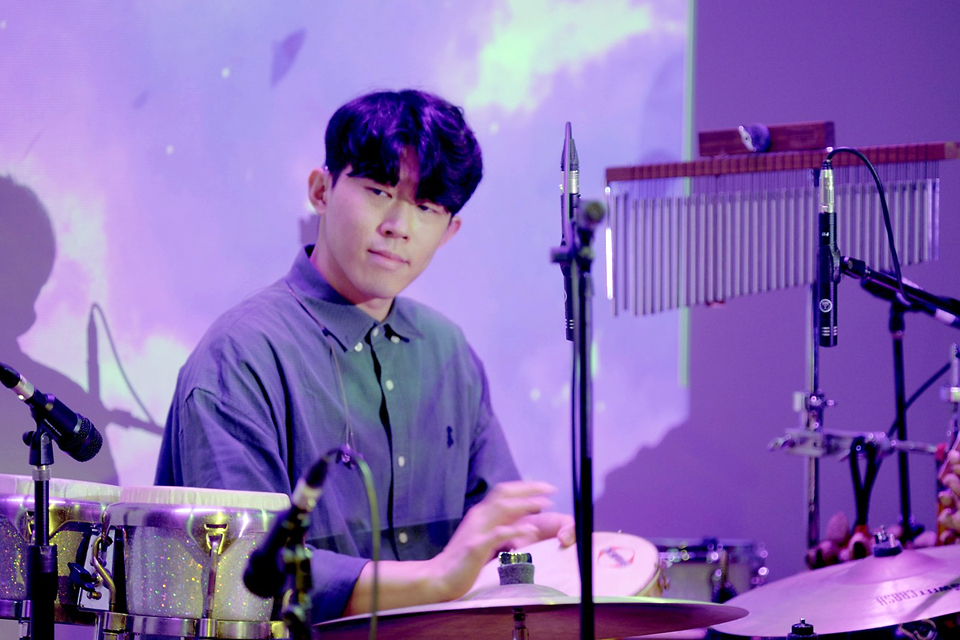 보라색 조명 아래에 콩가와 심벌, 윈드 차임 등의 악기가 놓여 있고 이성재 퍼커셔니스트가 손으로 악기를 연주하고 있다. 악기 주변에 여러 개의 마이크 스탠드가 설치되어 있다.