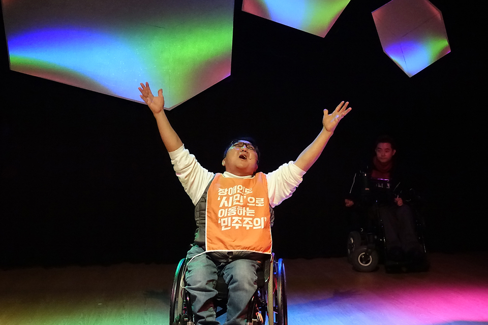 무대에서 휠체어를 탄 배우가 두 팔을 크게 들어 올리며 절규하고 있다. 배우는 “장애인도 ‘시민’으로 이동하는 ‘민주주의’라고 쓰인 주황색 슬로건 가리개를 몸에 두르고 있다. 뒤편 스크린 오브제가 무지갯빛으로 프로젝션되고 있다.