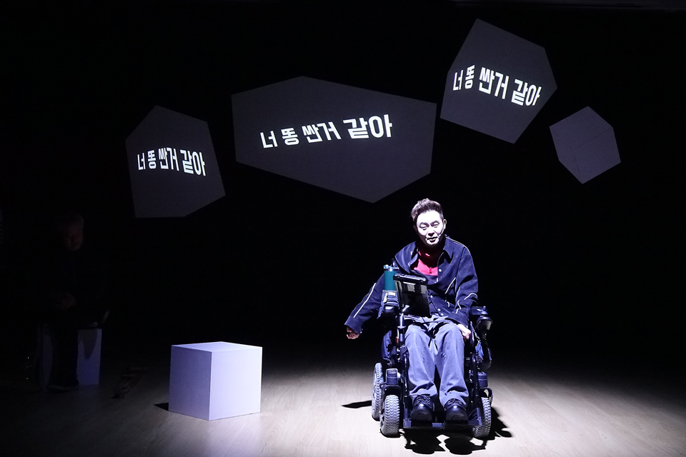 어두운 무대에서 핀조명 아래 휠체어를 탄 임일주 배우가 정면을 향해 앉아 있다. 뒷면 영상 자막 겸 오브제에는 “너 똥 싼 거 같아”라는 문장 세 개가 쓰여 있다.