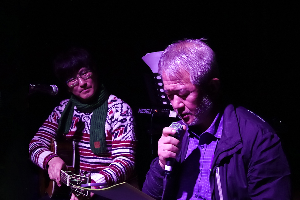 어두운 무대에서 두 명이 나란히 앉아 있다. 흰머리의 배우는 손에 든 마이크를 입 가까이 대고 두 눈을 감고 노래 부르고, 옆에서 악사가 기타를 치며 배우를 바라보고 있다.