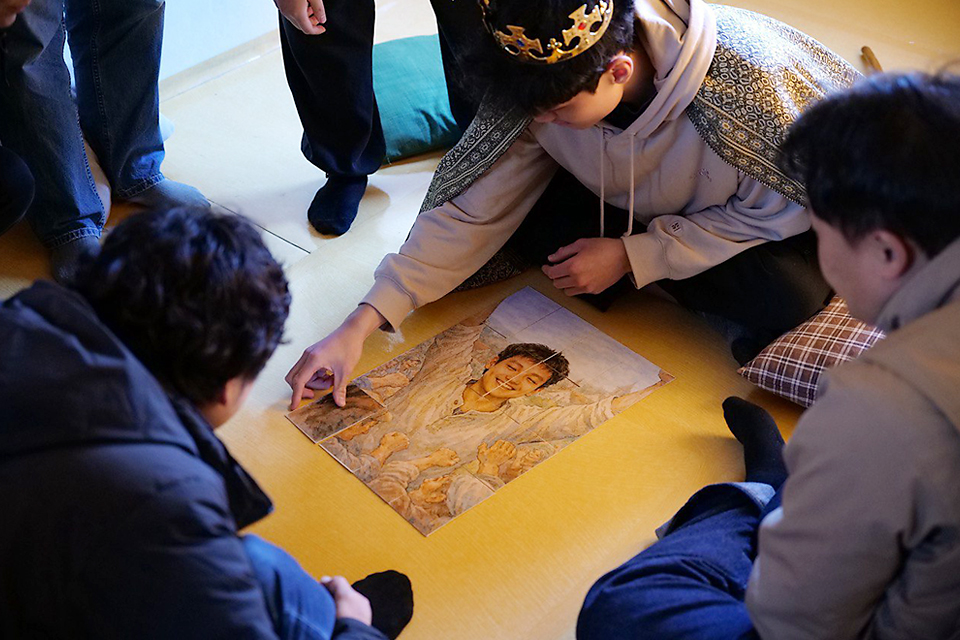 왕 역의 상일이 방바닥에 놓인 자기 모습의 퍼즐 그림카드의 마지막 장을 놓고 있고, 관객이 그 주변으로 모여 바라보고 있다. 