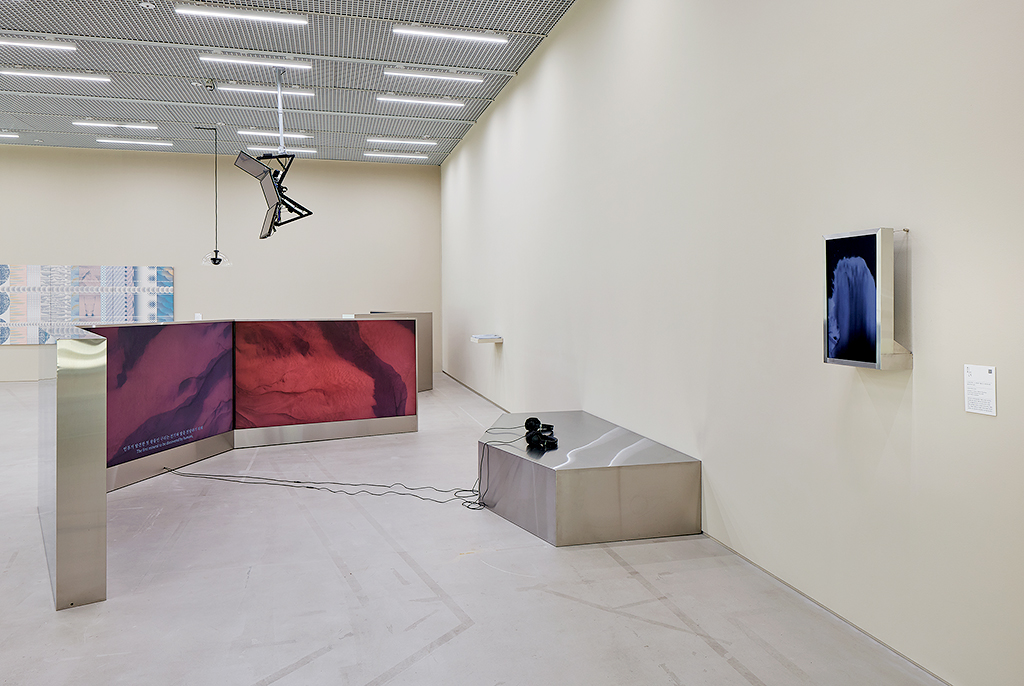 벽에 메탈 프레임의 작은 모니터가 걸려 있다. 바닥에 벽을 향해 메탈 프레임의 3채널 모니터가 나란히 연결되어 놓여 있고, 메탈 좌석에 유선 헤드폰이 놓여 있다. 영상에는 붉은 톤으로 확대한 땅의 이미지와 자막이 보인다.