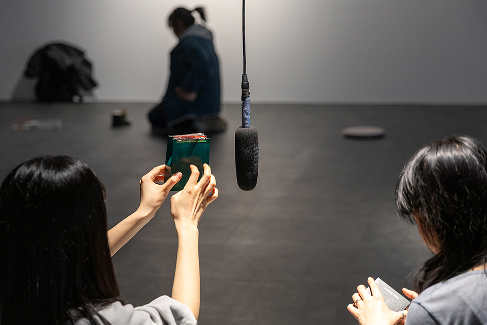 어두운 실내에서 두 여성이 마이크를 사이에 두고 등을 진 채 앉아 있다. 왼쪽 여성은 녹색 사각형 물건을 양손으로 들고 있으며, 오른쪽 여성은 회색 직사각형 물건을 오른손에 쥐고 마이크를 향해 있다. 배경에는 앉아 있는 또 다른 사람의 실루엣이 흐릿하게 보인다.