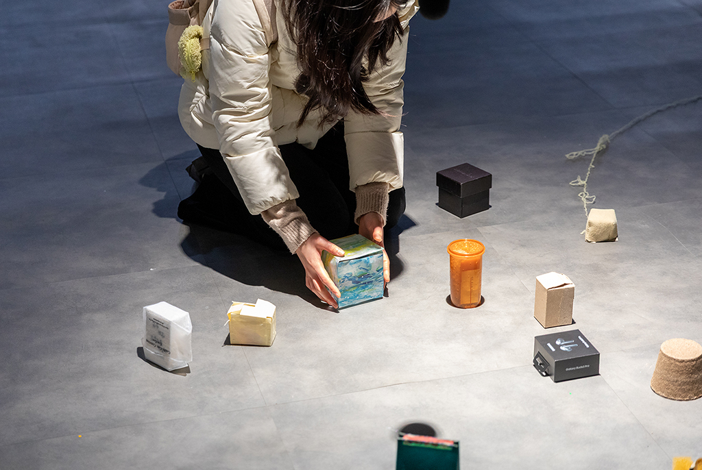 어두운 회색 바닥에 아이보리색 패딩을 입은 여성이 무릎을 꿇고 앉아, 푸른색 그림이 그려진 작은 상자를 양손으로 잡고 있다. 바닥에는 다양한 크기와 색상의 상자, 주황색 양초, 작은 화분, 털실 뭉치 등 여러 물건이 놓여 있다.