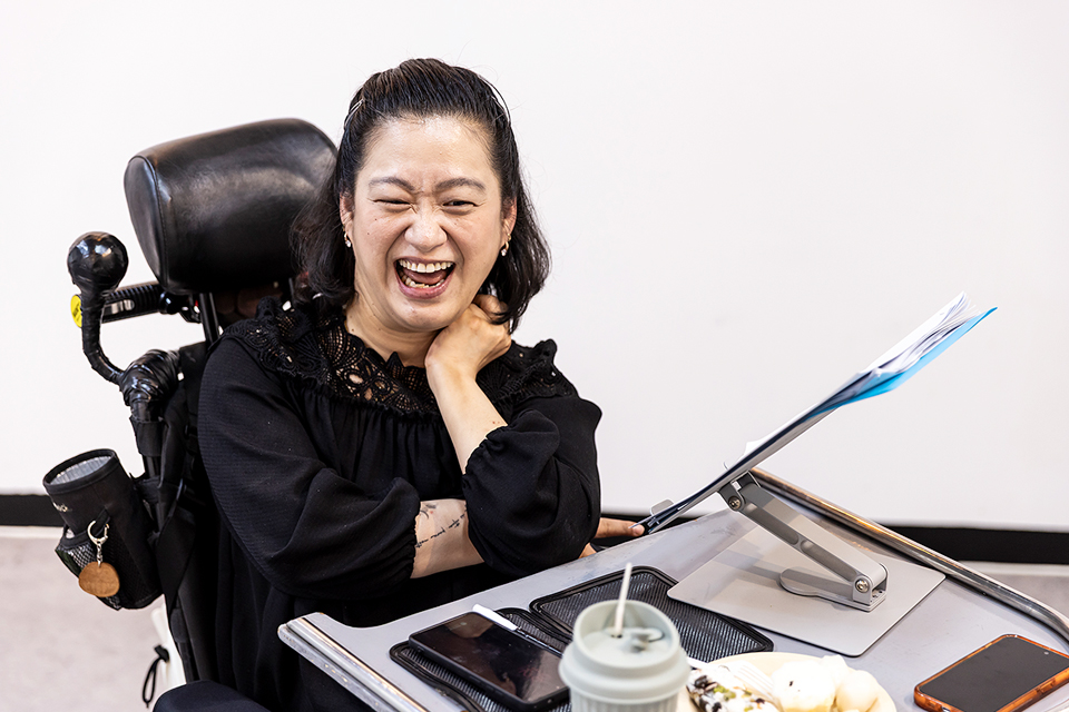 조우리 배우가 전동휠체어에 앉아 입을 크게 벌리며 웃고 있다. 트레이 위의 거치대에는 종이들이 올려져 있고, 그 옆에 음료컵과 스마트폰 등이 놓여 있다.