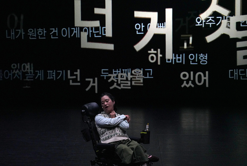 전동휠체어를 탄 조우리 배우가 어두운 무대 하단 중앙에 앉아 두 팔을 교차해 어깨를 감싸고 있다. 배경의 검은 화면에는 “넌 거짓말을 하고 있어”, “일어서자”, “내가 원한 건 이게 아닌데”, “비틀어져 놓게 펴기” 등 다양한 크기의 한글 텍스트가 흩어져 있다. 전동휠체어 옆에는 작은 음료병이 놓여 있다.