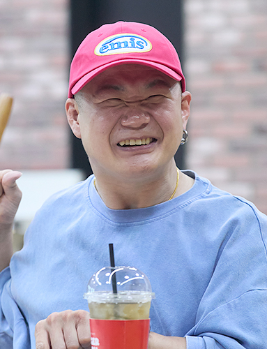 빨간 볼캡을 쓰고 한쪽에 귀고리를 한 파란 티셔츠를 입은 남성이 노트북을 앞에 두고 옆을 보며 웃고 있다.
