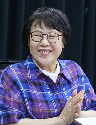 숏커트 머리에 뿔테 안경을 쓰고 파란 체크무늬 셔츠를 입은 여성이 두 손을 모으고 옆을 보며 웃고 있다. 