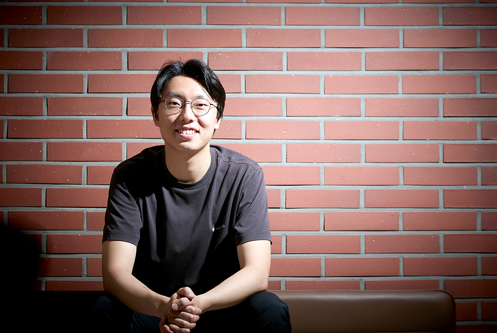 붉은 벽돌 벽을 배경으로 젊은 남성이 환하게 미소 지으며 두 손을 모으고 앉아 있다. 동그란 안경을 쓰고 검정색 반팔 티셔츠를 입고 있다.
