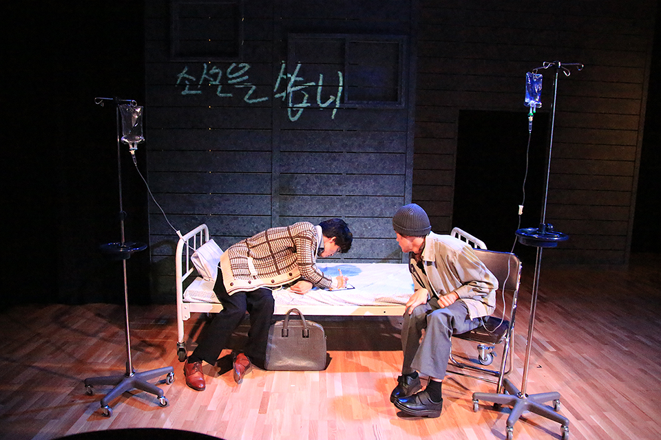 벽면에 ‘소설을 씁니다’라는 문구가 새겨진 무대 배경 아래 침대가 놓여 있다. 침대 위에는 한 배우가 숙여 무언가를 쓰고 있고, 그 오른쪽 옆 의자에 앉은 배우가 그 모습을 바라보고 있다. 침대 양옆으로는 수액 거치대가 서 있다.